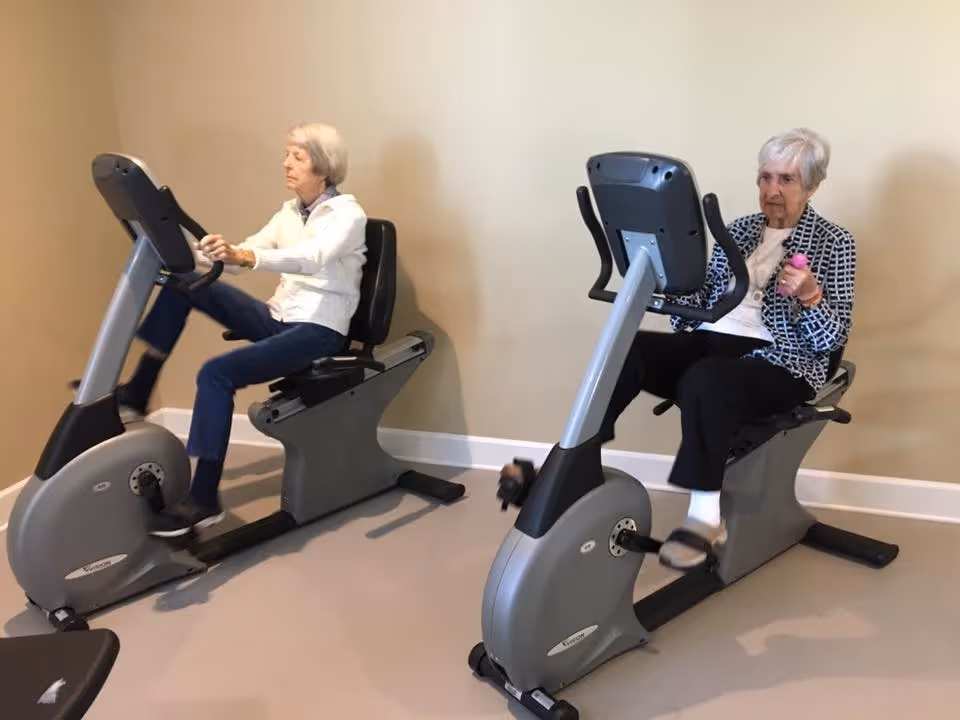 Two elderly women exercising on recumbent stationary bikes in a fitness room with beige walls and carpeted floor.