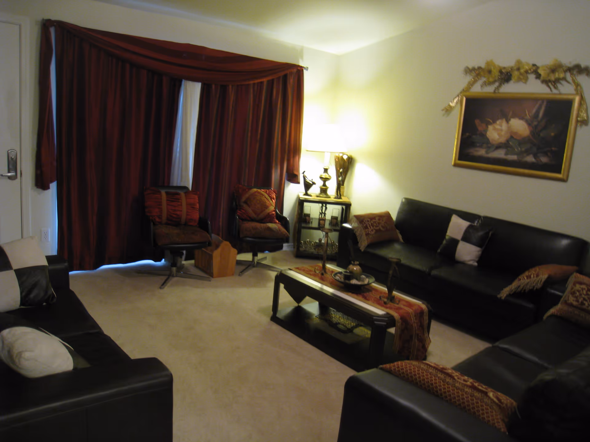 Living room with dark leather sofas around a central coffee table, two chairs by closed red curtains, a side table lamp, and wall art.
