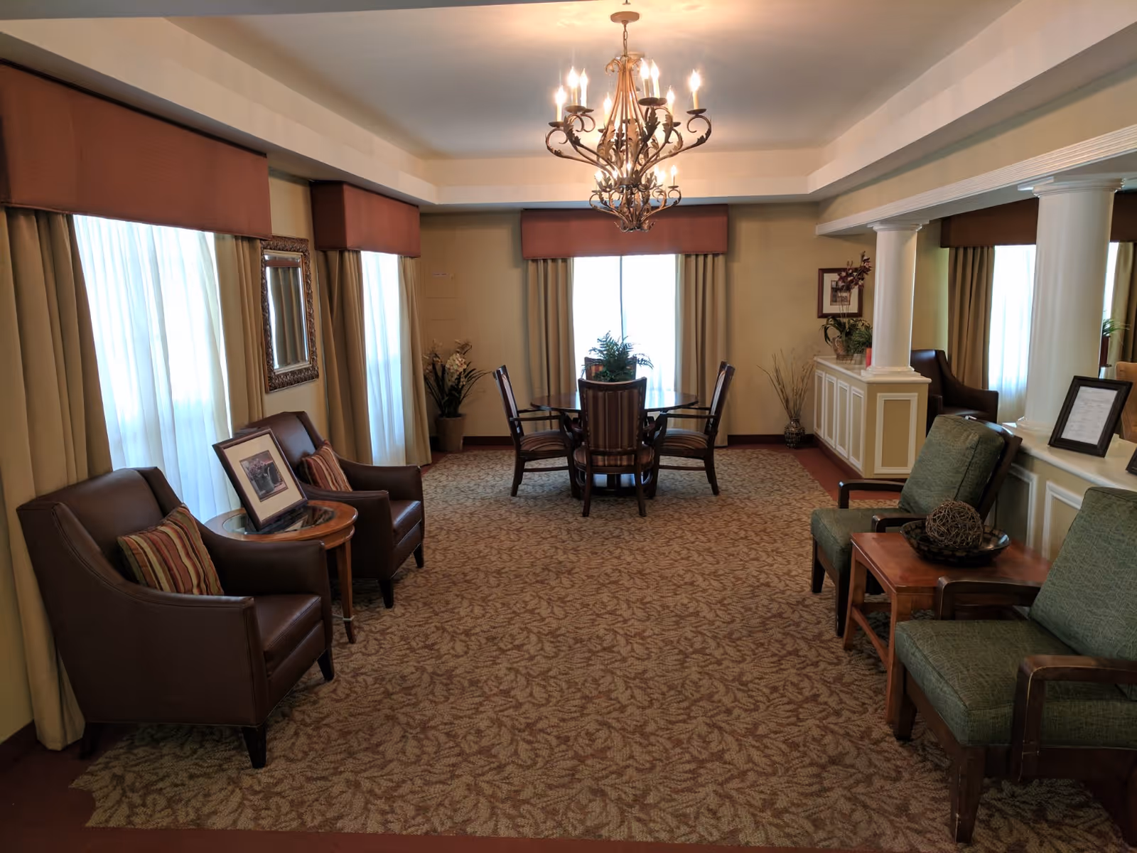 A cozy sitting area with multiple chairs arranged around small tables, featuring a chandelier hanging from the ceiling. The room has large windows with curtains and valances, decorative plants, framed pictures on the walls, and a patterned carpet covering the floor.