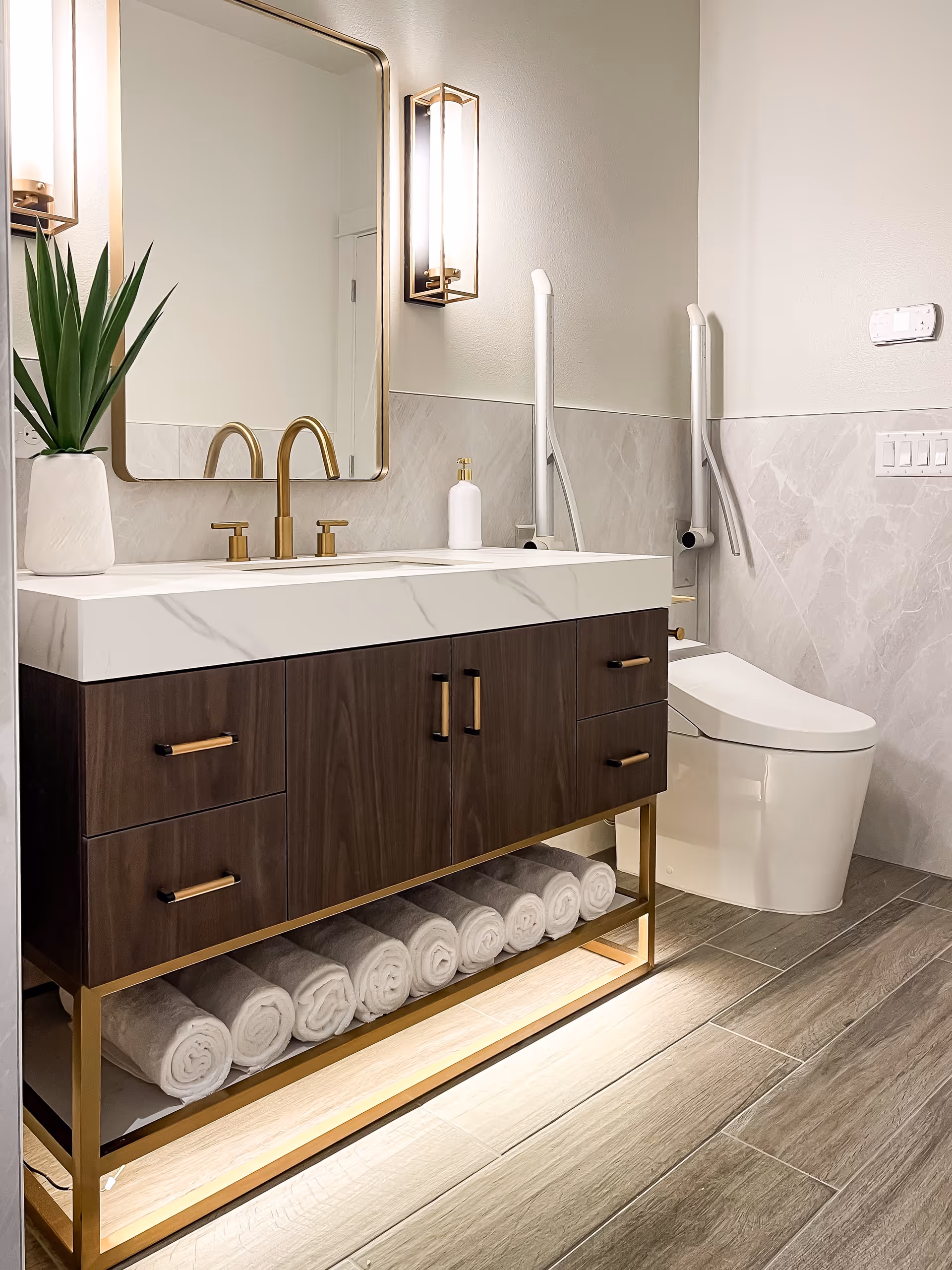Modern bathroom with a wooden vanity featuring a white marble countertop and gold fixtures. A large rectangular mirror with a gold frame is mounted above the sink, flanked by two vertical wall lights. Below the vanity, several rolled white towels are neatly arranged. To the right, there is a white toilet with safety grab bars installed on the wall. The floor is covered with wood-look tiles, and the walls have a light gray marble tile halfway up.