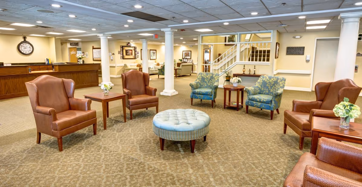 A spacious senior living facility common area with multiple armchairs arranged around a central round ottoman. The room features beige walls, carpeted floors, white columns, and a reception desk in the background. There are framed pictures on the walls and a staircase leading to an upper level.
