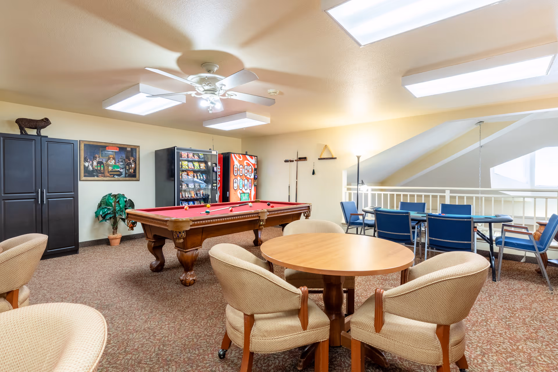 A recreation room featuring a billiards table, vending machines, and seating areas with chairs and tables. The room has bright lighting and a cozy atmosphere.