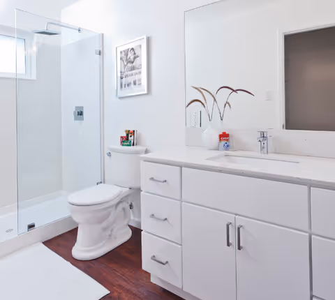 A clean and modern bathroom featuring a glass-enclosed shower, a white toilet, and a white vanity with a sink and countertop. On the countertop, there is a white vase with decorative dried plants and a container of Colgate toothpaste. A large mirror is mounted above the vanity, and a framed black and white picture hangs on the wall above the toilet. The floor has a dark wood finish with a white bath mat in front of the shower.