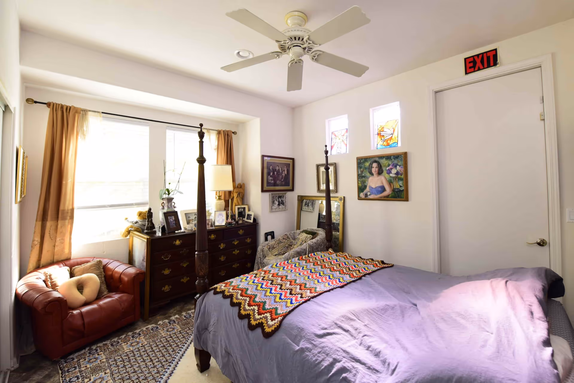 A bright bedroom with a four-poster bed covered with a purple bedspread and a colorful crocheted blanket. The room has a red leather armchair with a beige pillow, a wooden dresser with framed photos and a lamp, and several paintings and mirrors on the walls. Two small stained glass windows are above a white door with an exit sign. A ceiling fan is mounted on the white ceiling.