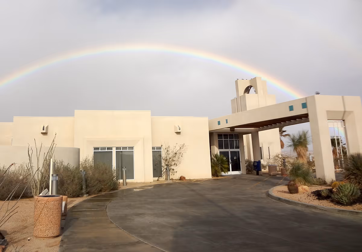 The front entrance of a low-rise beige building with a driveway and a vivid rainbow arching overhead.