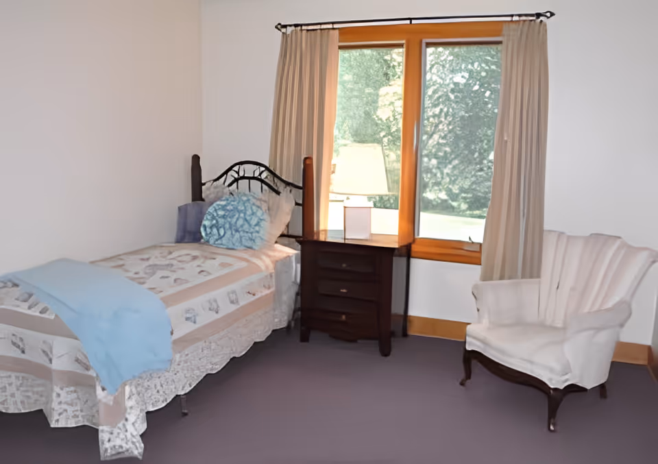 Sunlit bedroom with a single bed, bedside table and lamp by a window and an upholstered armchair.