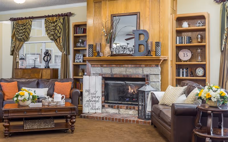 Cozy living room with two brown leather sofas adorned with orange and beige pillows, a wooden coffee table with a flower arrangement and tea set, a stone fireplace with a wooden mantel decorated with vases, candles, and a large letter B, built-in wooden bookshelves filled with books and decorative items, and windows with patterned curtains.