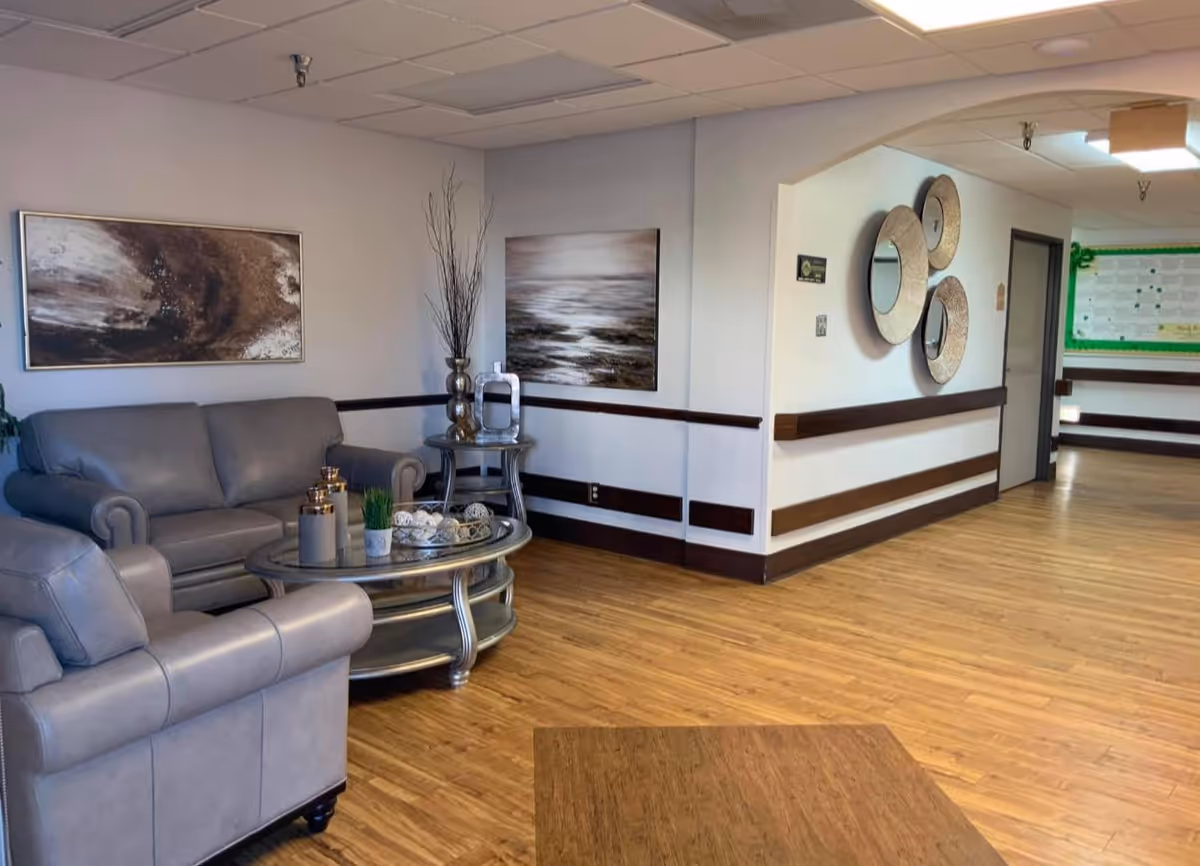 A cozy seating area in a nursing and rehabilitation facility with two gray leather armchairs and a matching sofa arranged around a glass coffee table. The walls are decorated with abstract paintings and circular mirrors. The floor is wooden, and the space is well-lit with ceiling lights.