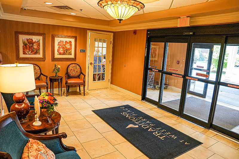 Senior living facility lobby with seating, decorative paintings, a side table with flowers and lamp, and automatic glass entrance doors with a floor mat.