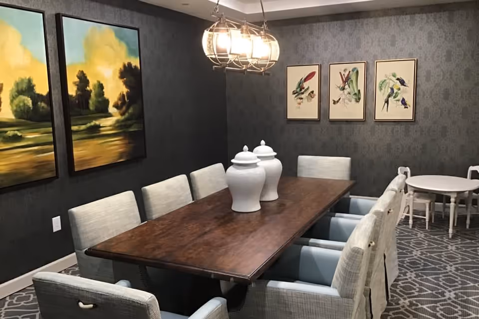 Elegant dining room with a long wooden table surrounded by upholstered chairs, decorative white vases, framed artwork and a chandelier.