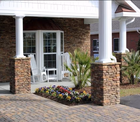 Covered front porch with stone pillars, white rocking chairs, and a flowerbed in front of an entrance.