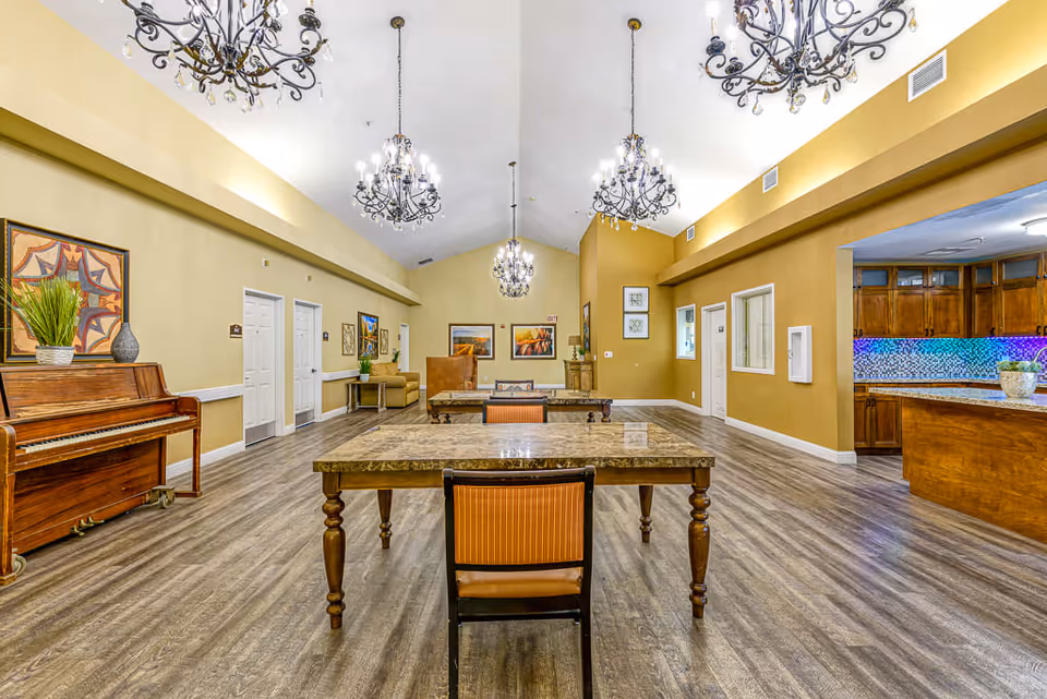 Interior view of a senior living facility common area with wooden tables and chairs, a piano on the left, chandeliers hanging from a high ceiling, and a kitchen area with wooden cabinets and a colorful backsplash on the right.