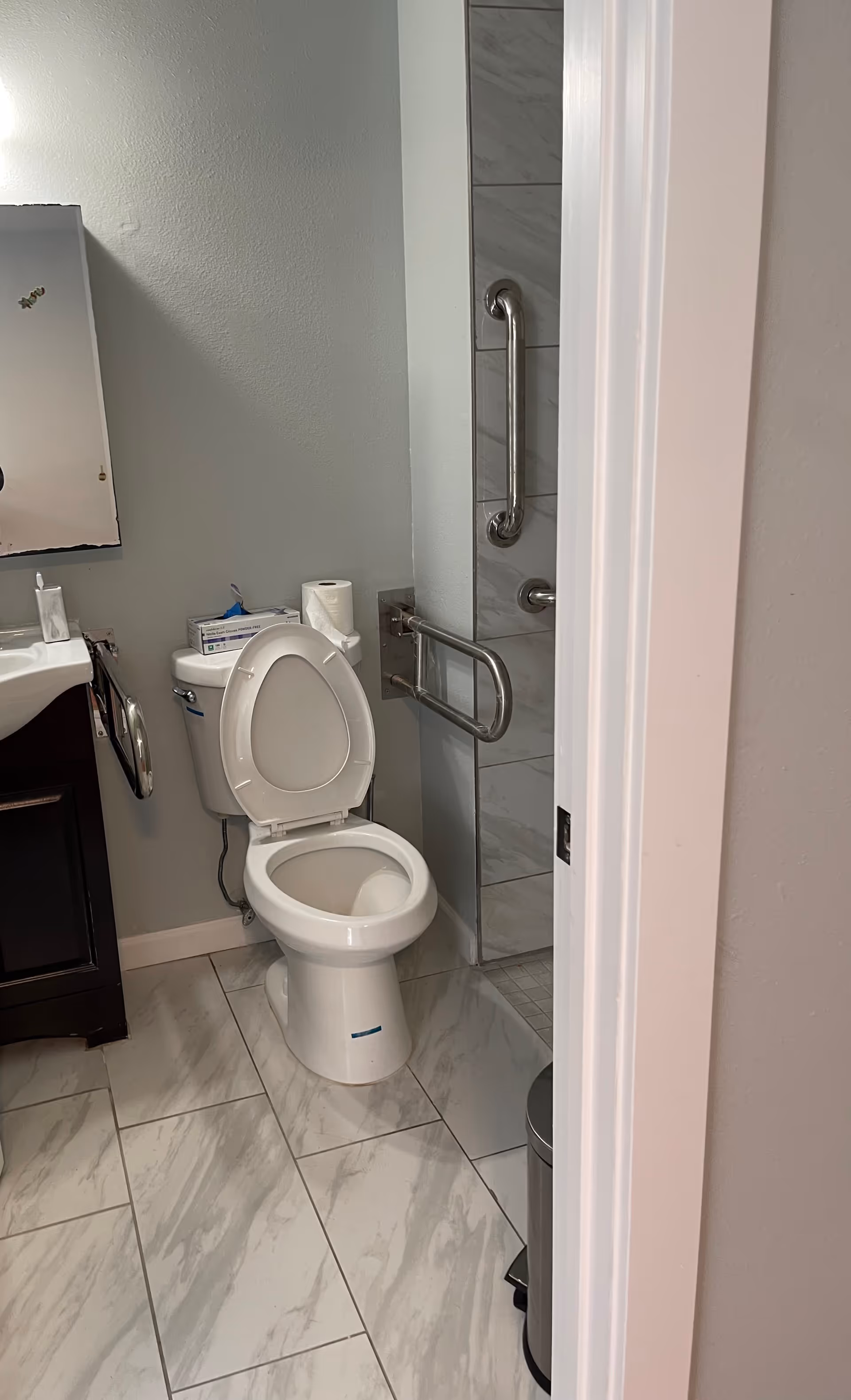 Accessible bathroom showing a toilet with raised seat, grab bars, tiled floor, and a partial sink and mirror.
