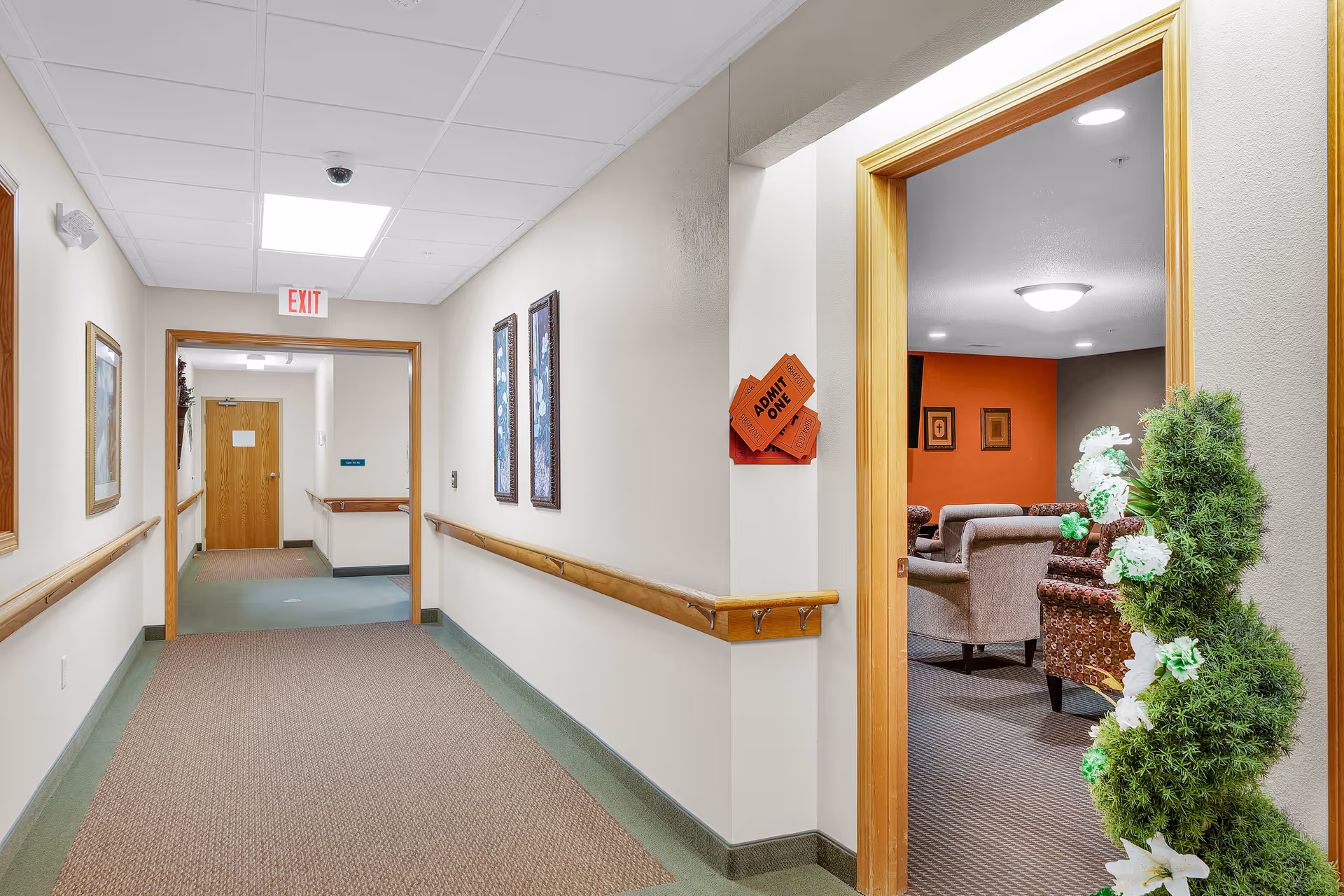 A well-lit hallway in an assisted living facility with beige walls, wooden handrails, and carpeted floors. The hallway leads to a wooden door with an exit sign above it. On the right side, there is an open doorway showing a cozy lounge area with armchairs, orange and brown walls, framed pictures, and a decorative green plant with white flowers near the entrance.