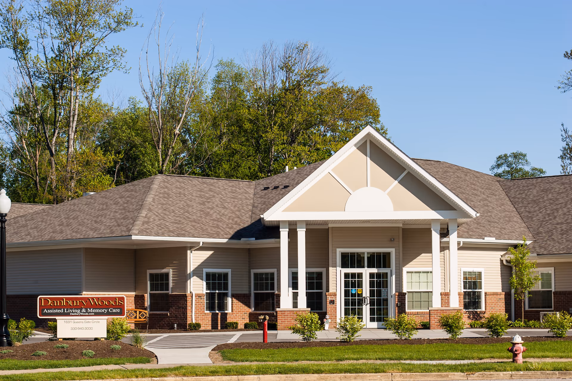 Exterior front view of Danbury Woods Assisted Living & Memory Care facility with a beige and brick facade, white columns, and a peaked roof. The building is surrounded by green grass, small bushes, and trees under a clear blue sky.