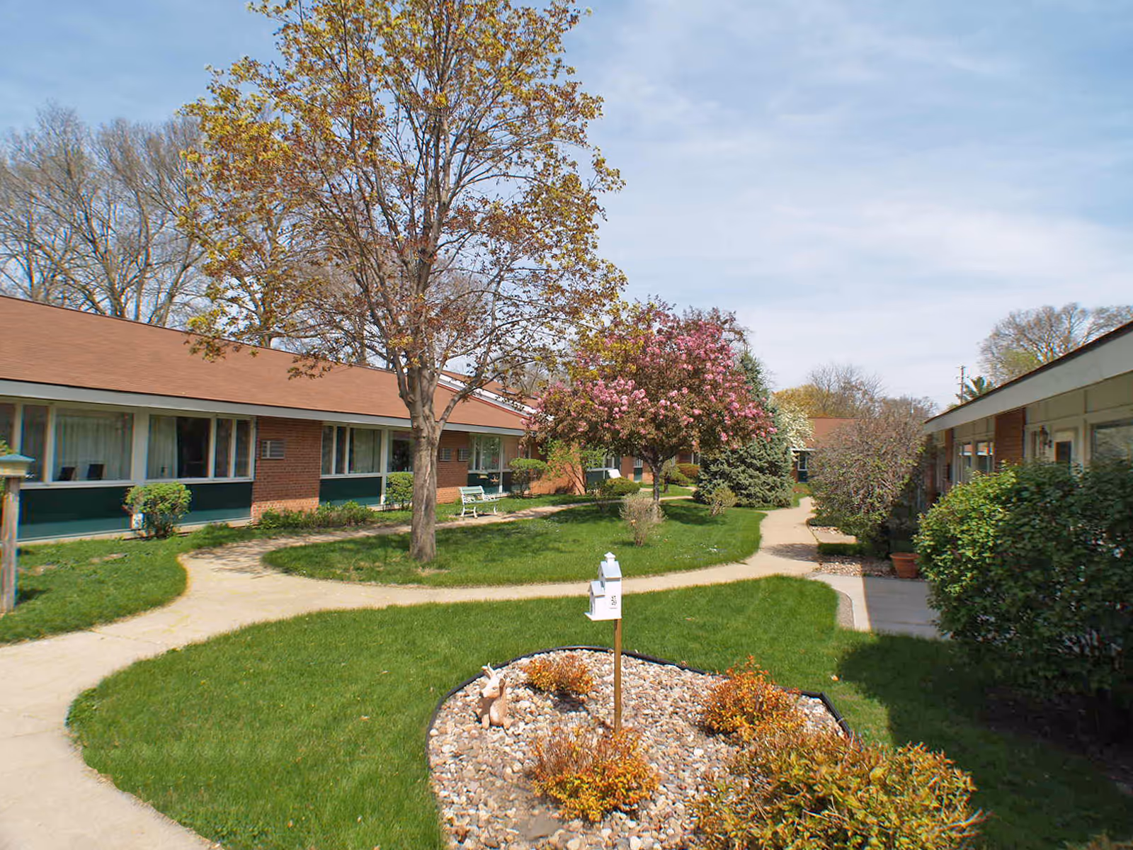 Grass courtyard with winding walkways, trees, flowering shrubs and single-story brick assisted living buildings surrounding it.