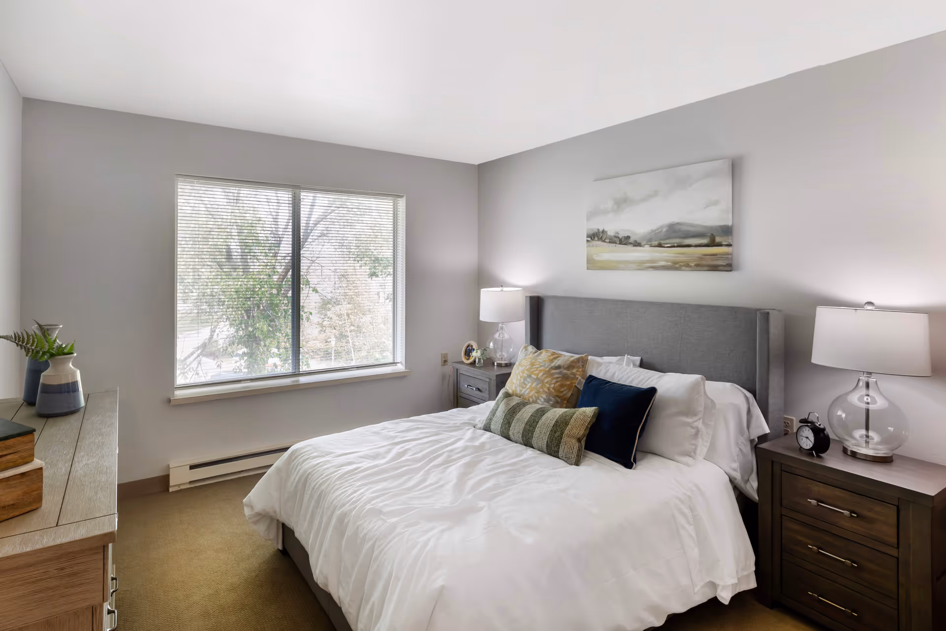A neatly made bed with white bedding and multiple decorative pillows in a bedroom. The room has a large window with blinds, two bedside tables with lamps, an alarm clock, and a piece of landscape artwork hanging above the bed. There is also a dresser with decorative items on the left side of the room.