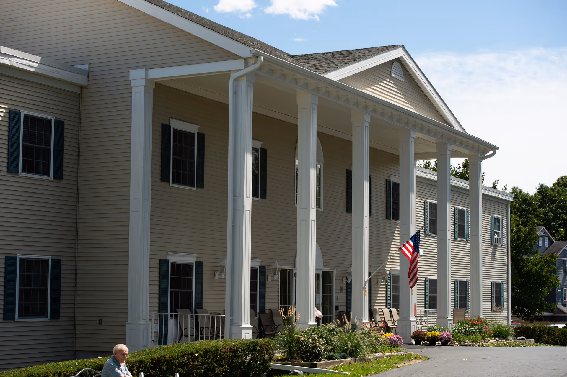 Front exterior of a two-story beige assisted living building with tall white columns, an American flag, landscaping, and a person seated near the entrance.