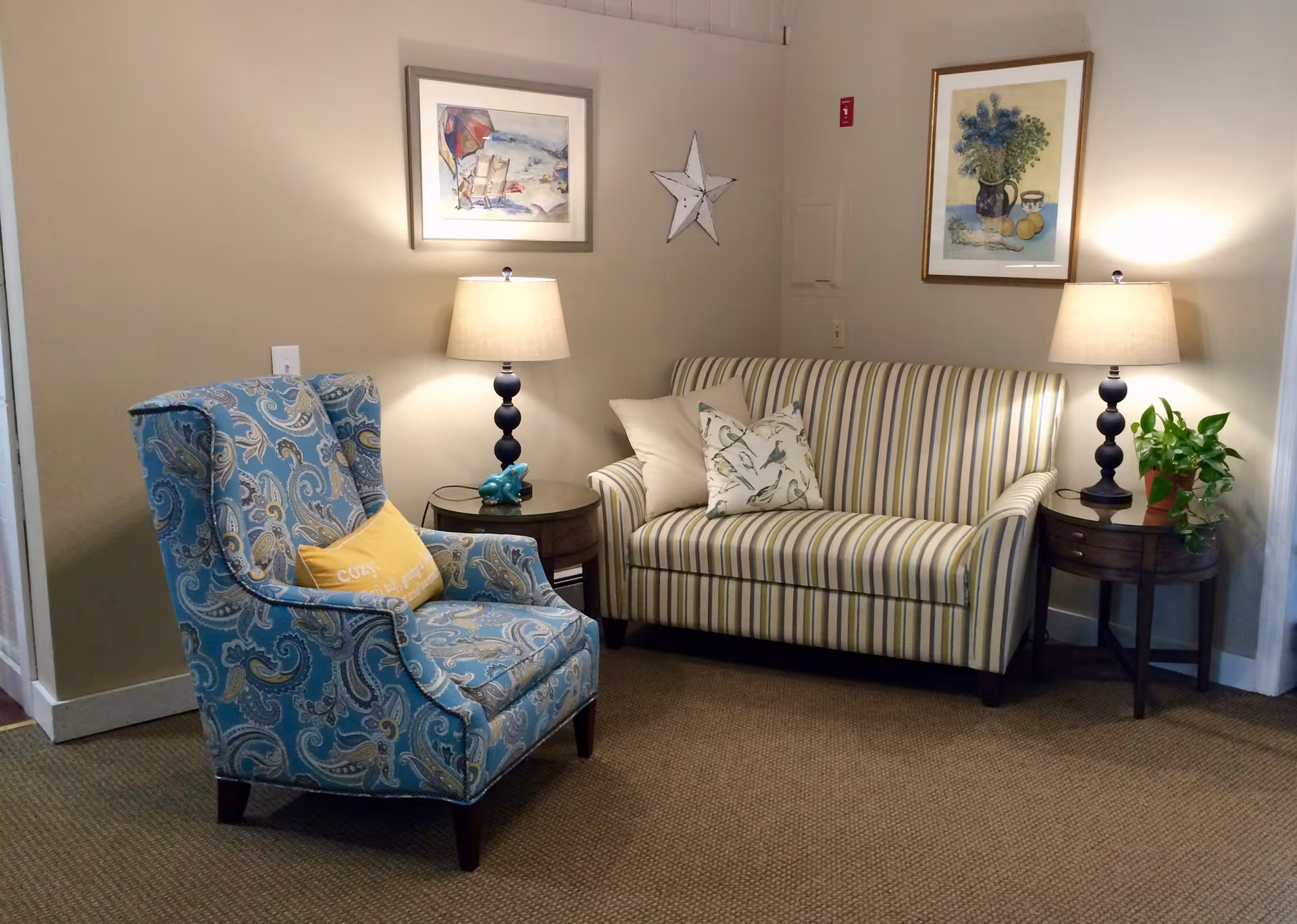 A cozy sitting area with a blue patterned armchair featuring a yellow pillow, a striped loveseat with two decorative pillows, two side tables each holding a lamp, a small plant on one table, and framed artwork on the beige walls.