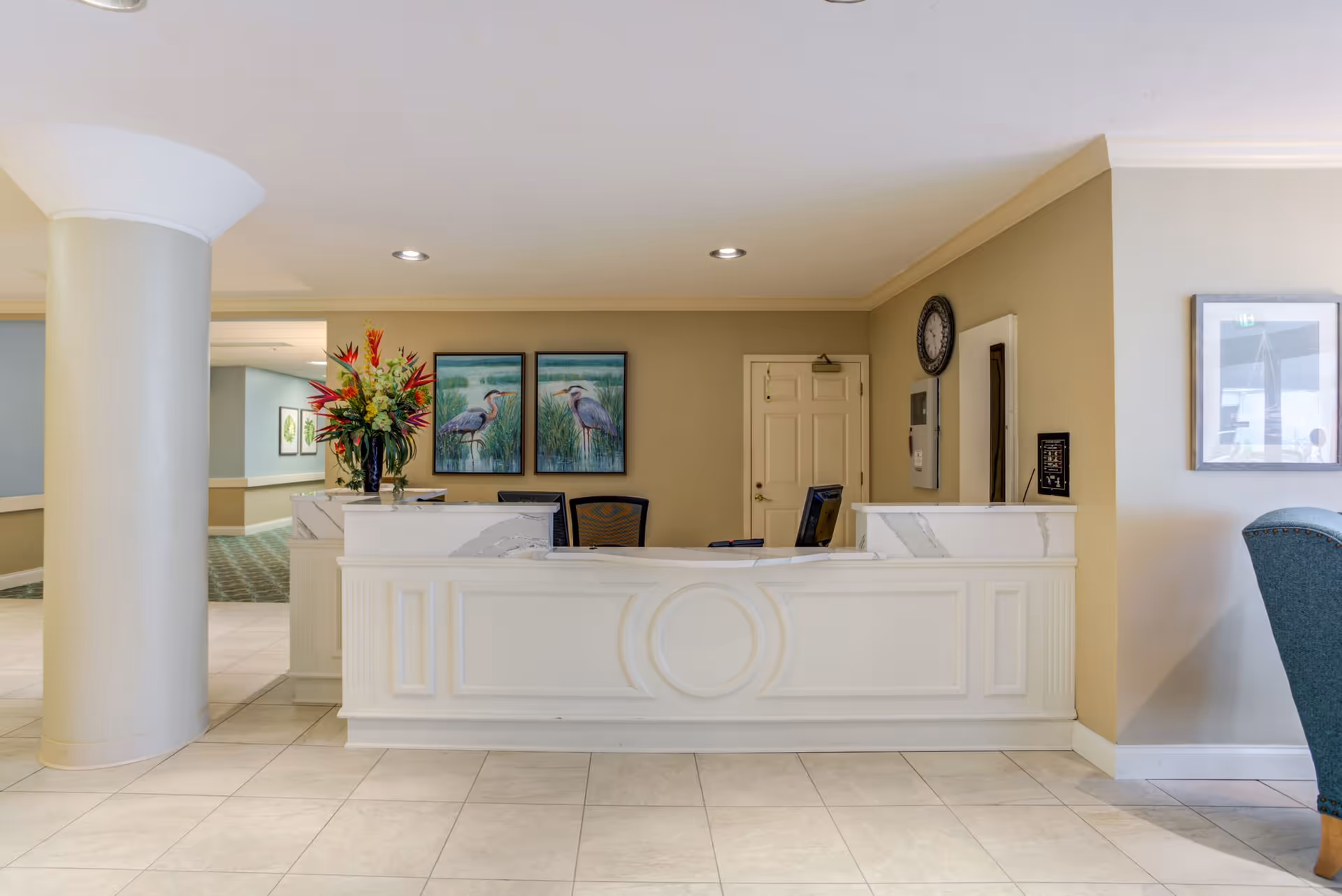 Reception desk area in a senior living facility with a white counter, two computer monitors, a large flower arrangement, and two framed paintings of herons on the wall behind. There is a large round column to the left and a blue upholstered chair partially visible on the right.
