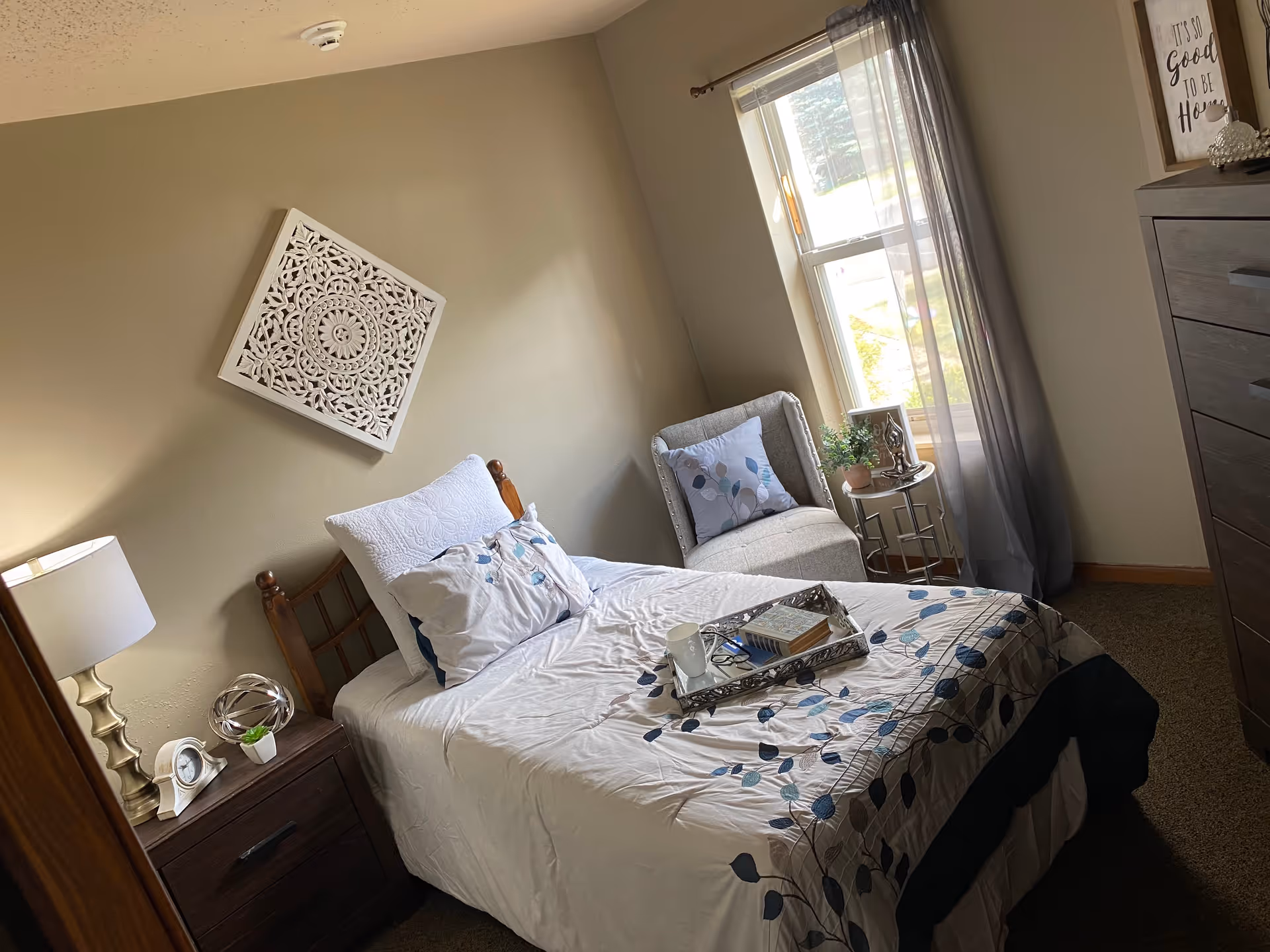 A cozy bedroom with a single bed made up with white and blue floral bedding. There is a wooden nightstand with a lamp, a clock, and a small plant. A decorative white carved wall art hangs above the bed. Next to the window is a cushioned chair with a pillow, a small round table with a plant and decorative items. A dresser is visible on the right side of the image. The window has sheer gray curtains allowing natural light to enter the room.