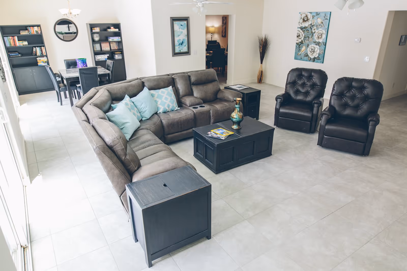 A spacious living room with a large gray sectional sofa adorned with light blue and patterned throw pillows, two black leather armchairs, a black coffee table with decorative items, and a side table. In the background, there is a dining area with a table and chairs, bookshelves, and wall art. The room has light-colored tiled flooring and white walls.