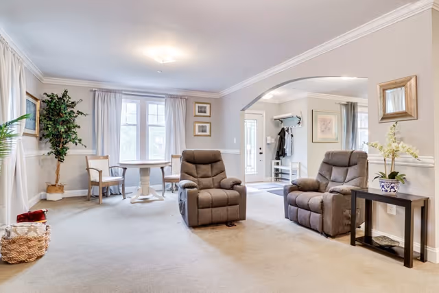 A bright and spacious living room in Cobbdale Assisted Living featuring two gray recliner chairs, a small black side table with a potted orchid, a round wooden table with two chairs near a window with white curtains, a large potted plant, and an entryway with coat hooks and a bench visible through an archway.