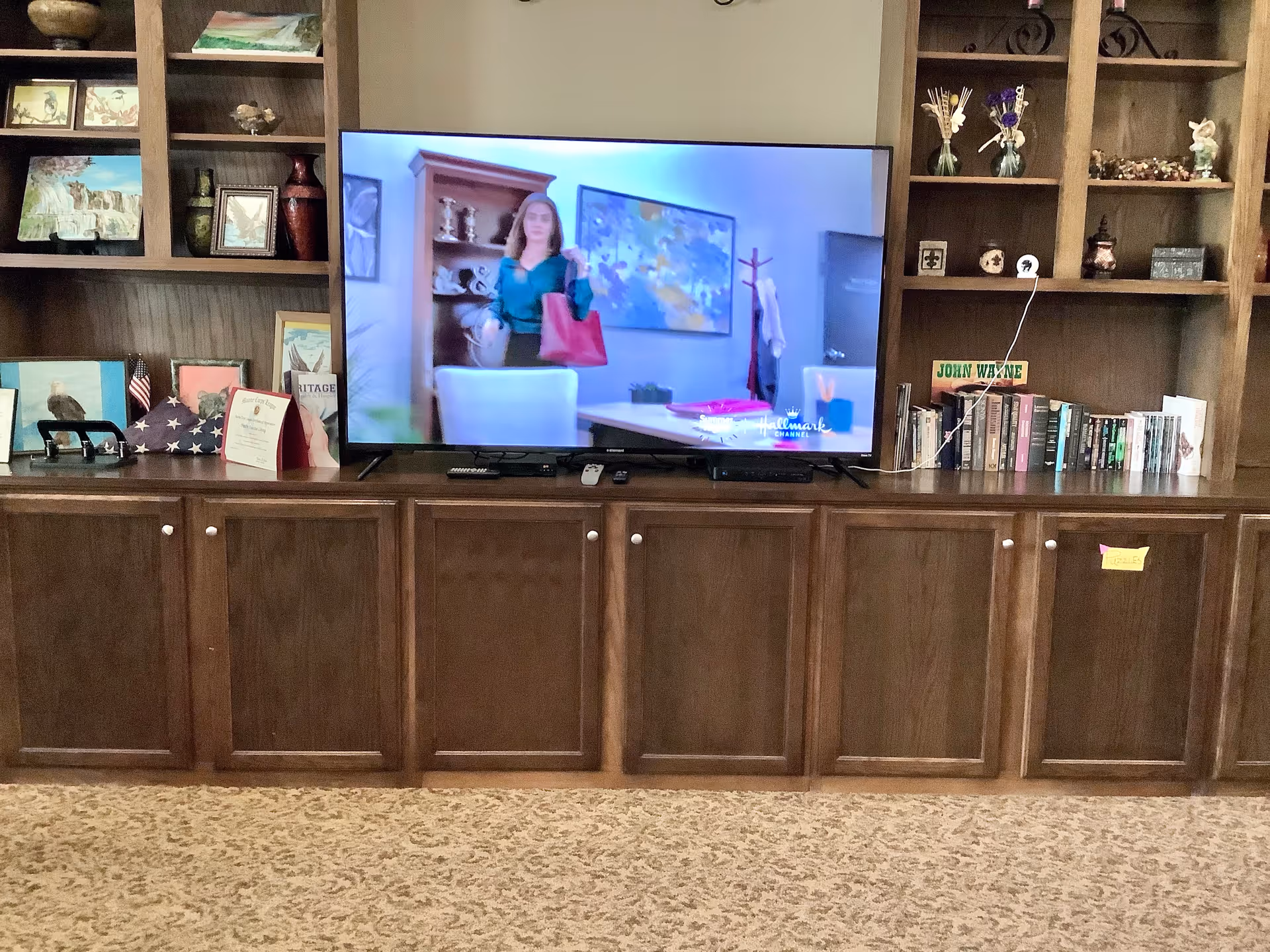 A large flat-screen TV is placed on a wooden cabinet with multiple closed storage compartments. The TV is displaying a scene with a woman holding a red bag. Surrounding the TV are built-in wooden shelves filled with various decorative items, framed pictures, books, and small vases. The floor is carpeted with a patterned design.