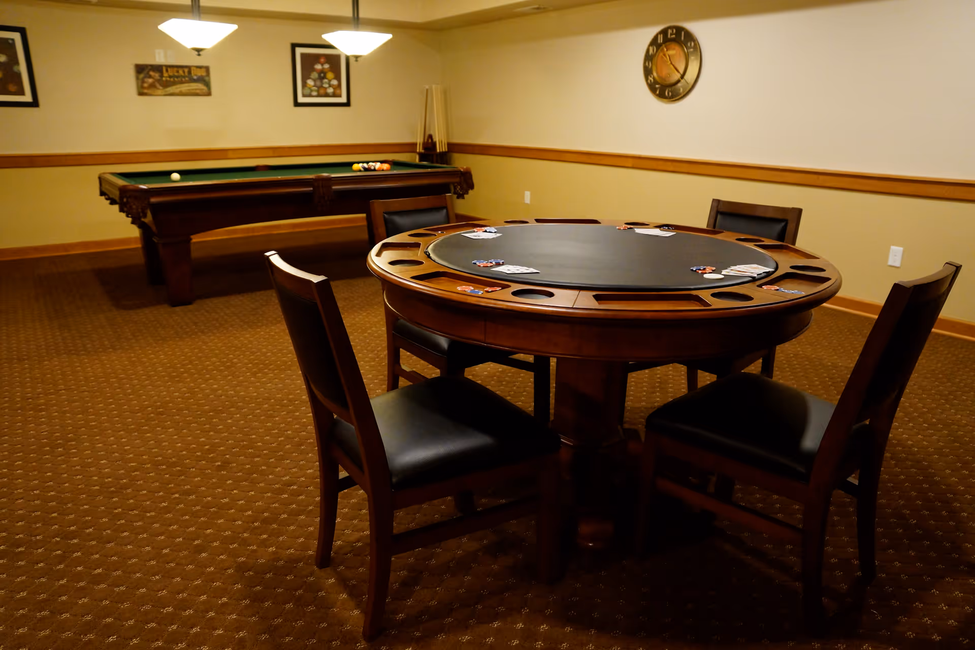 A game room with a round poker table surrounded by four chairs, poker chips and playing cards on the table, and a pool table in the background under two hanging lights. The room has carpeted flooring, beige walls with wooden trim, framed pictures, and a wall clock.
