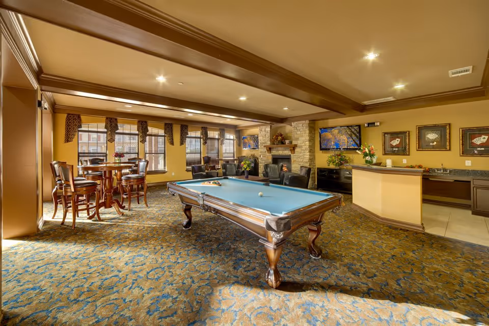 A spacious recreational room with a pool table in the center, surrounded by comfortable seating including armchairs and high chairs around a tall table. The room features large windows with patterned valances, a stone fireplace, two wall-mounted televisions, and a small kitchenette area with a counter and sink. The carpet has a blue and brown pattern, and the walls are painted in warm tones.