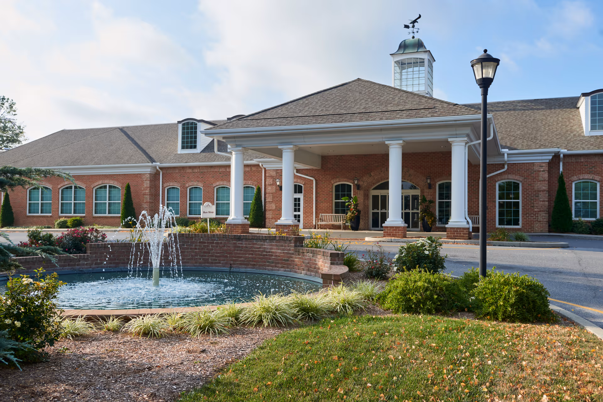 Exterior view of a brick building with white columns at the entrance, a cupola with a weather vane on the roof, a water fountain in front, landscaped bushes and grass, and a street lamp near the driveway.