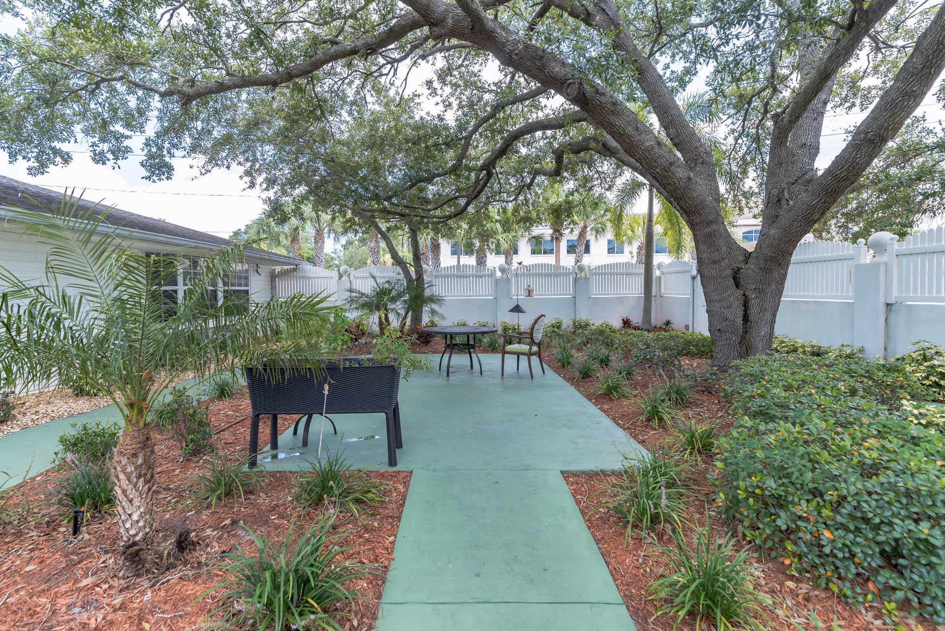 Outdoor garden area with a large tree providing shade over a green concrete patio. There is a black bench and a round table with two chairs on the patio. The area is surrounded by plants, shrubs, and a white fence in the background.
