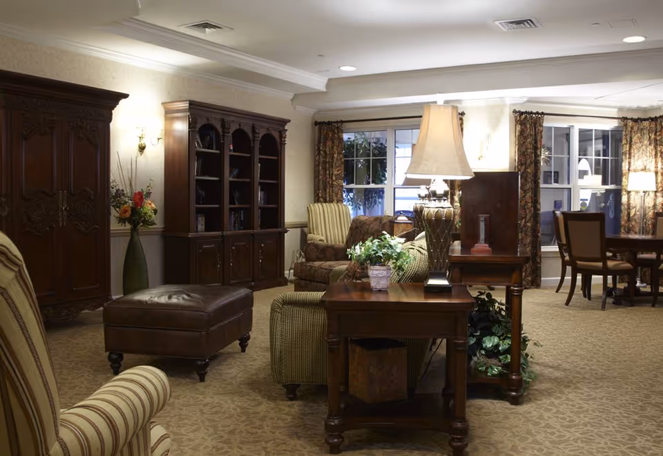 A cozy living room area in a senior living facility featuring upholstered armchairs, a leather ottoman, wooden side tables with lamps, a large wooden cabinet, a bookshelf, floral curtains, and a carpeted floor.