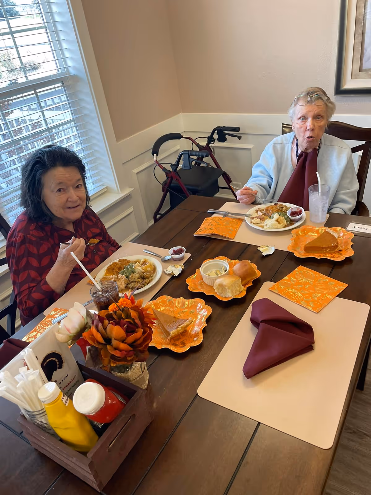 Two elderly women sitting at a dining table in a well-lit room, enjoying a meal with plates of food including mashed potatoes, rolls, and pie. One woman is wearing a red patterned top and the other a light blue sweater with a maroon napkin tucked in. A walker is positioned behind the woman in the blue sweater. The table is set with orange decorative plates and napkins, and there is a small flower arrangement and condiments on the table.
