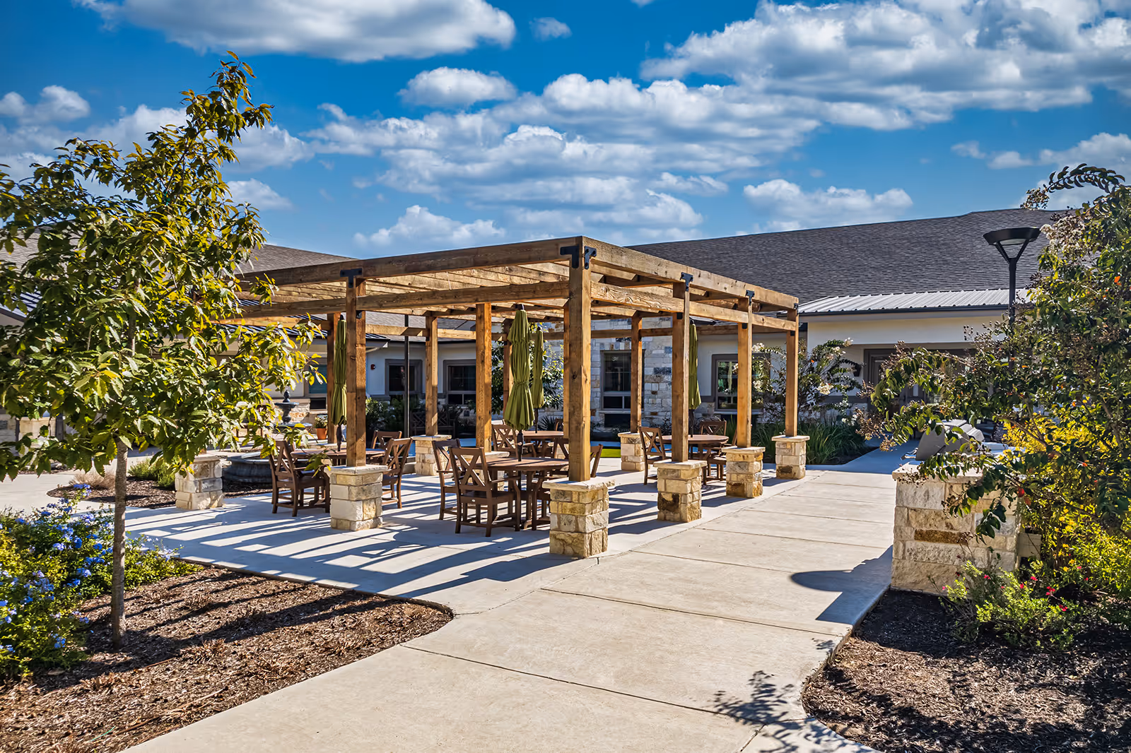 Outdoor patio area at Volante Senior Living of Sage Spring featuring a wooden pergola with stone pillars, several tables and chairs underneath, surrounded by landscaped plants and trees under a partly cloudy blue sky.