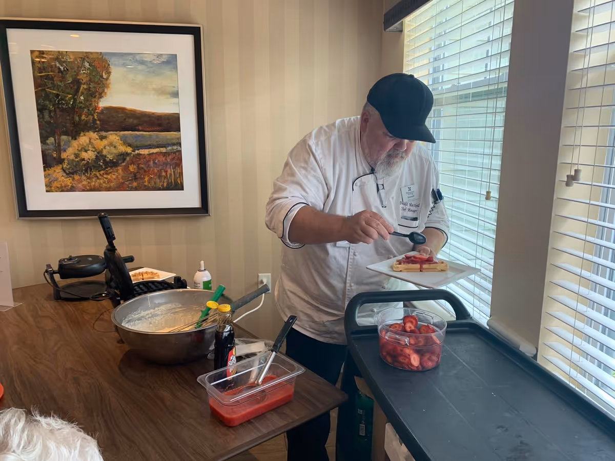 A chef wearing a white uniform and black cap is preparing a plate of waffles topped with strawberries in a kitchen area. On the counter, there is a large mixing bowl with batter, a bottle of syrup, a container of sliced strawberries, and a waffle maker. A framed landscape painting hangs on the wall behind the chef.