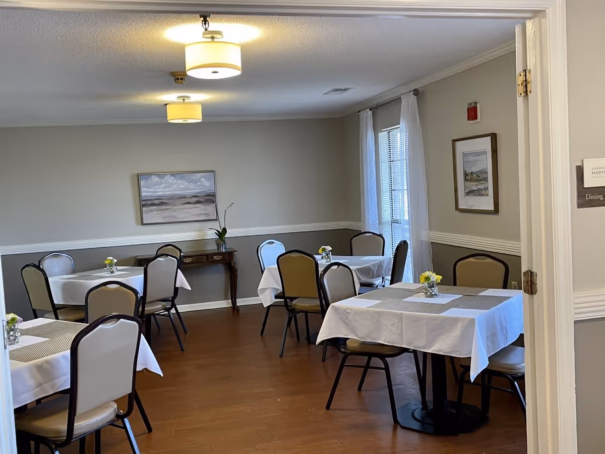 A dining room with several tables covered with white tablecloths and beige placemats. Each table has chairs around it and small flower arrangements as centerpieces. The room has light gray walls with white trim, two ceiling lights, framed artwork on the walls, and a window with white curtains letting in natural light.