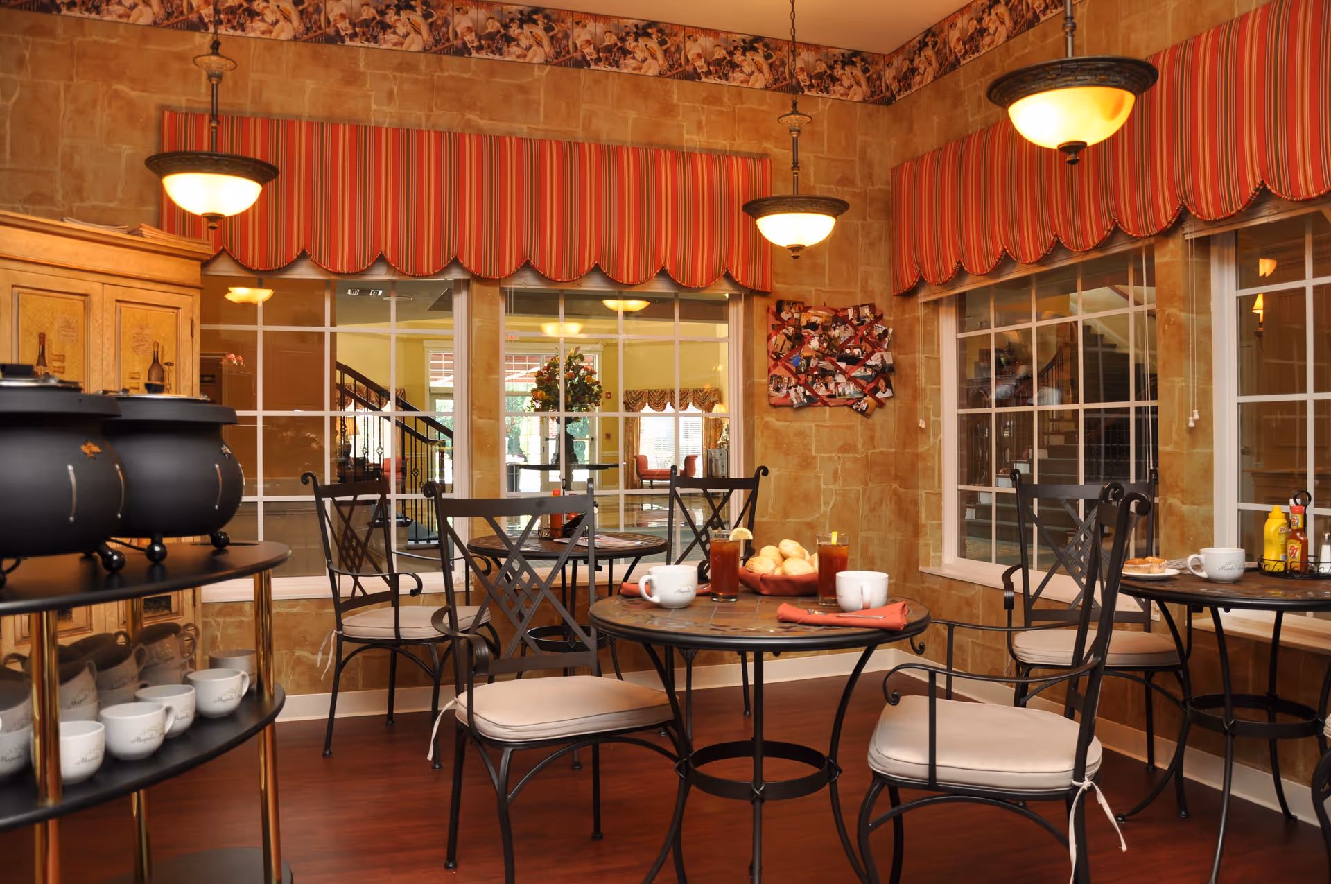 Cozy dining area with round iron tables and cushioned chairs, striped valances over large windows, and warm pendant lighting.