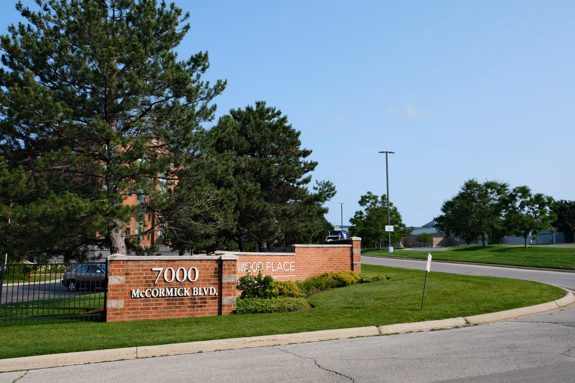 Brick entrance sign for Lincolnwood Place at 7000 McCormick Blvd, surrounded by green grass, bushes, and trees with a clear blue sky in the background.