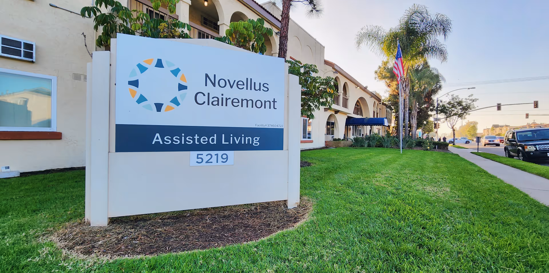 Outdoor view of Novellus Clairemont assisted living facility sign on a grassy area with the building and an American flag in the background along a sidewalk and street.
