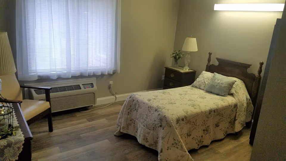 A small bedroom with a single bed covered in a floral bedspread and two pillows. There is a wooden nightstand with a lamp and a small plant next to the bed. A window with sheer white curtains lets in natural light. A chair and a table with a lace cover are partially visible on the left side of the room. The floor is wood laminate.