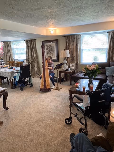 A cozy living room in an adult living facility with beige carpet and light-colored walls. There are two large windows with curtains letting in natural light. A woman is playing a harp in the center of the room. Several elderly residents are seated around the room, some in wheelchairs. The room is furnished with a brown leather sofa, wooden tables, a lamp, and floral decorations. A walker with a face mask hanging on it is visible in the foreground.