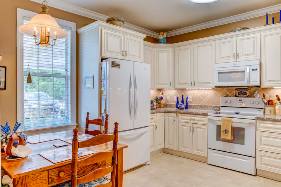 Bright kitchen with white cabinets, a white refrigerator, a white stove with a microwave above it, and a wooden dining table with two chairs near a window with blinds. The kitchen has beige tiled backsplash and under-cabinet lighting.