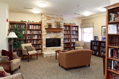 A cozy living room area with a stone fireplace centered between two tall bookshelves filled with books. The room has several upholstered chairs and a sofa arranged around a wooden coffee table. There is a window with a valance letting in natural light, and a potted plant is visible in the corner.