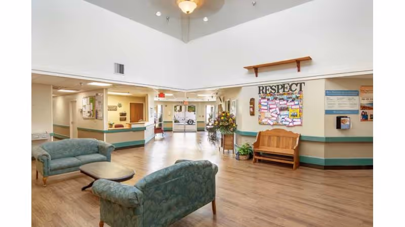 A spacious nursing home lobby with wooden flooring, two green upholstered sofas facing each other with a small wooden coffee table between them, a wooden bench against the wall, a bulletin board with the word RESPECT above it, and a flower arrangement on a small table. The area is well-lit with ceiling lights and has a wide entrance with glass doors at the far end.