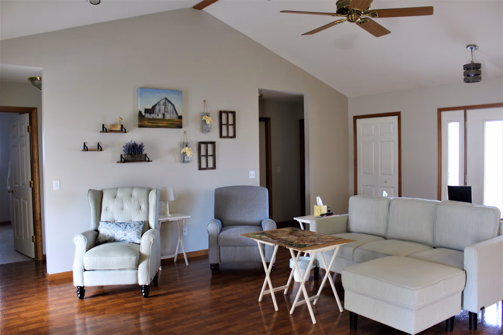 A bright living room with wooden flooring, featuring a light gray sectional sofa with a matching ottoman, a gray armchair, and a cream-colored tufted wingback chair with a decorative pillow. There are two small side tables with a lamp and tissue box. The walls are decorated with small shelves, a painting of a barn, and hanging flower jars. Ceiling fans with wooden blades are mounted on the vaulted ceiling. Doors and doorways lead to other rooms.