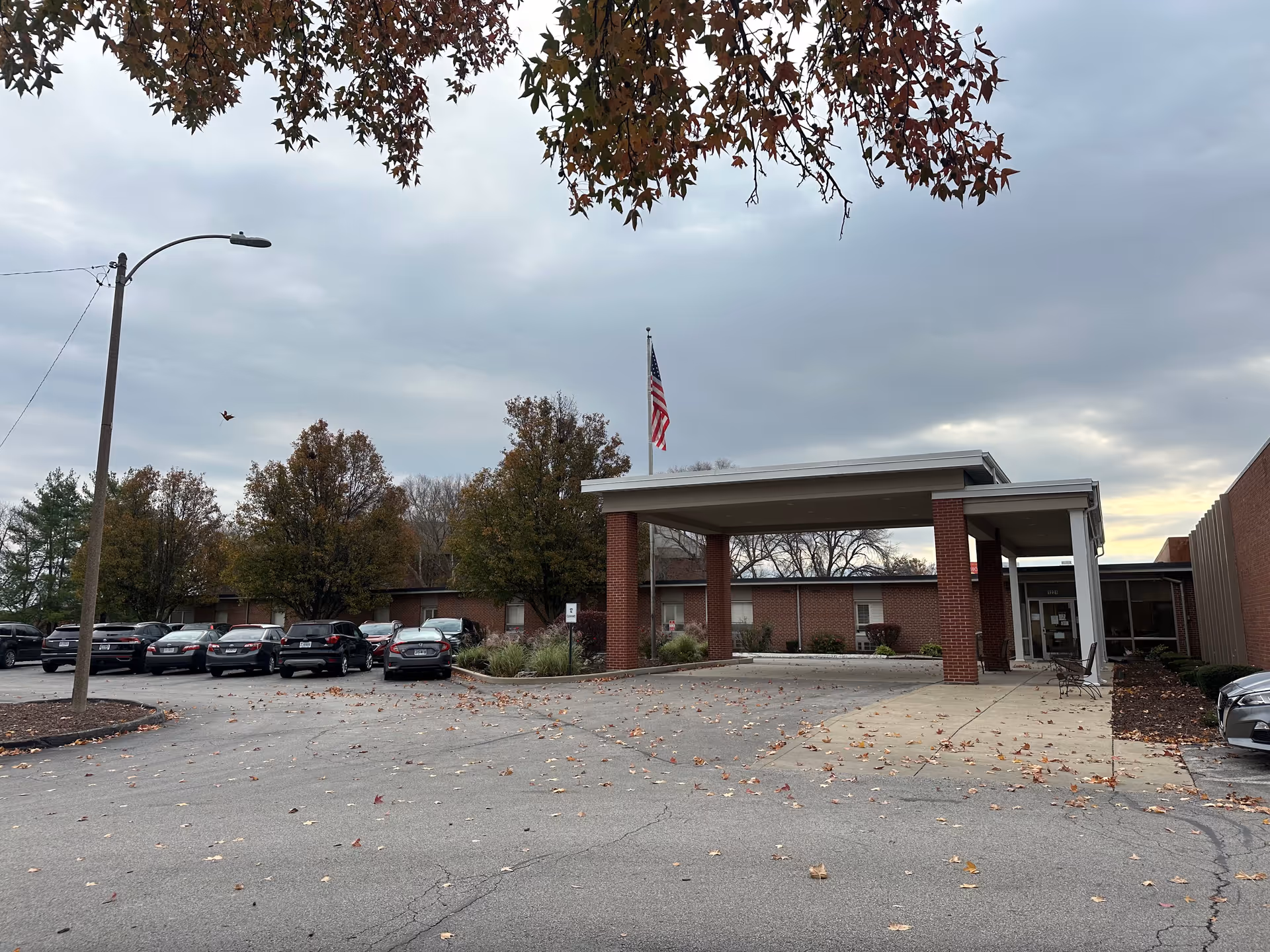 Exterior view of LEWIS and CLARK GARDENS facility showing a covered entrance with brick pillars, an American flag on a flagpole, parked cars, and trees with autumn leaves under a cloudy sky.