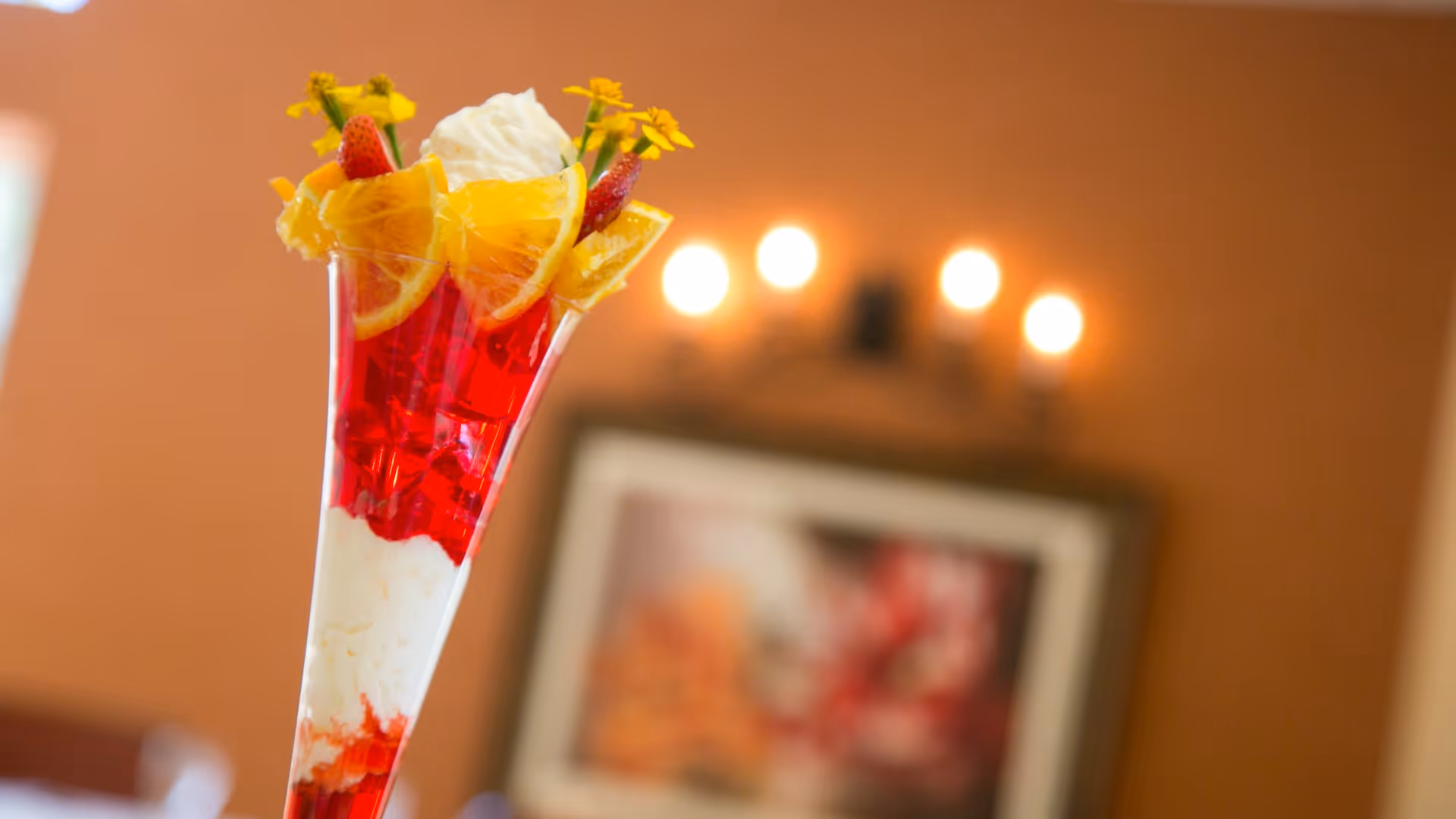 A tall dessert glass filled with a layered red gelatin and white creamy dessert, topped with orange slices, strawberries, whipped cream, and small yellow flowers, set against a blurred background with warm lighting and a framed picture on the wall.