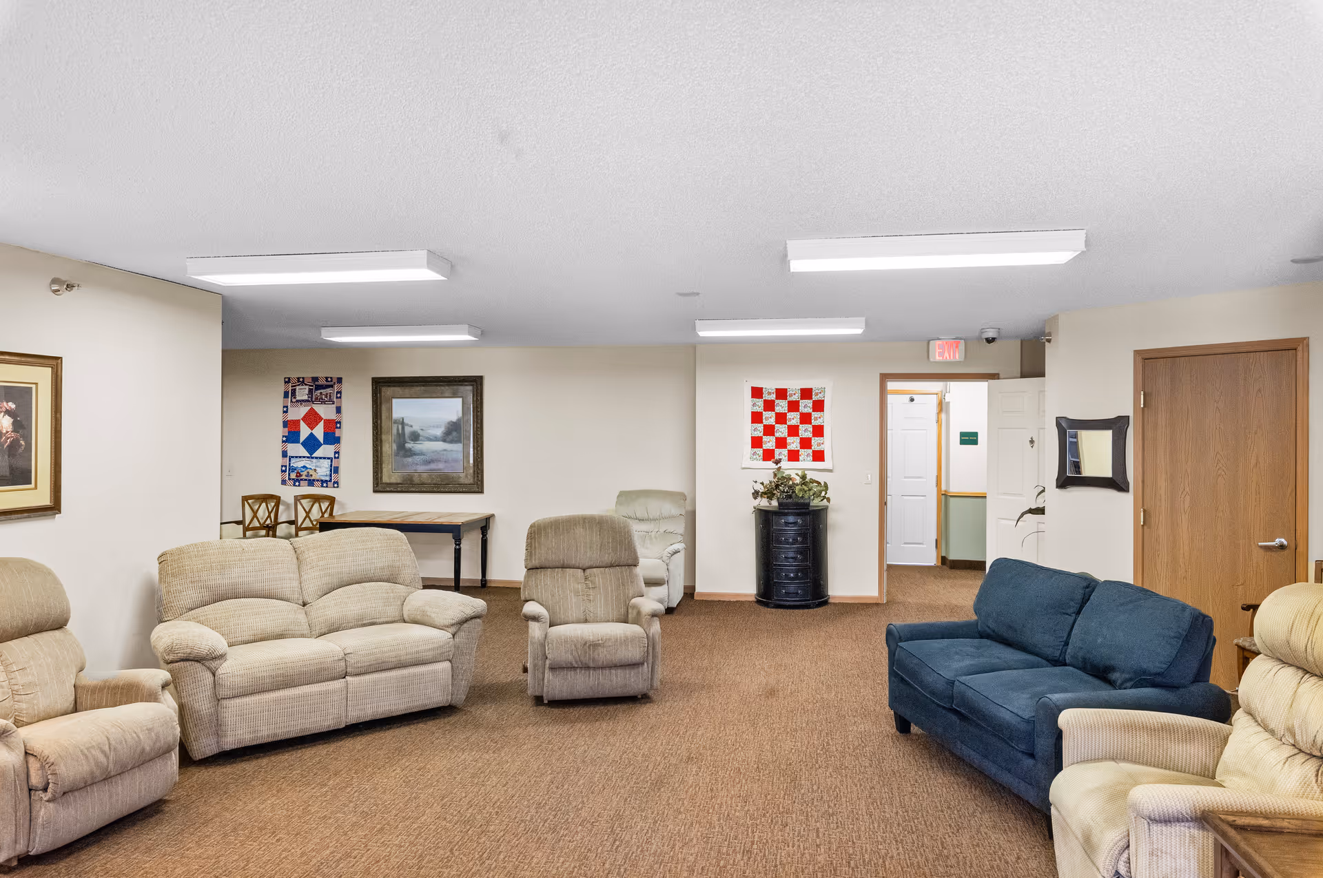 A senior living facility common area with multiple upholstered chairs and sofas arranged around the room. The walls are decorated with framed pictures and quilted wall hangings. There is a small table with chairs in the background and a door leading to another room.