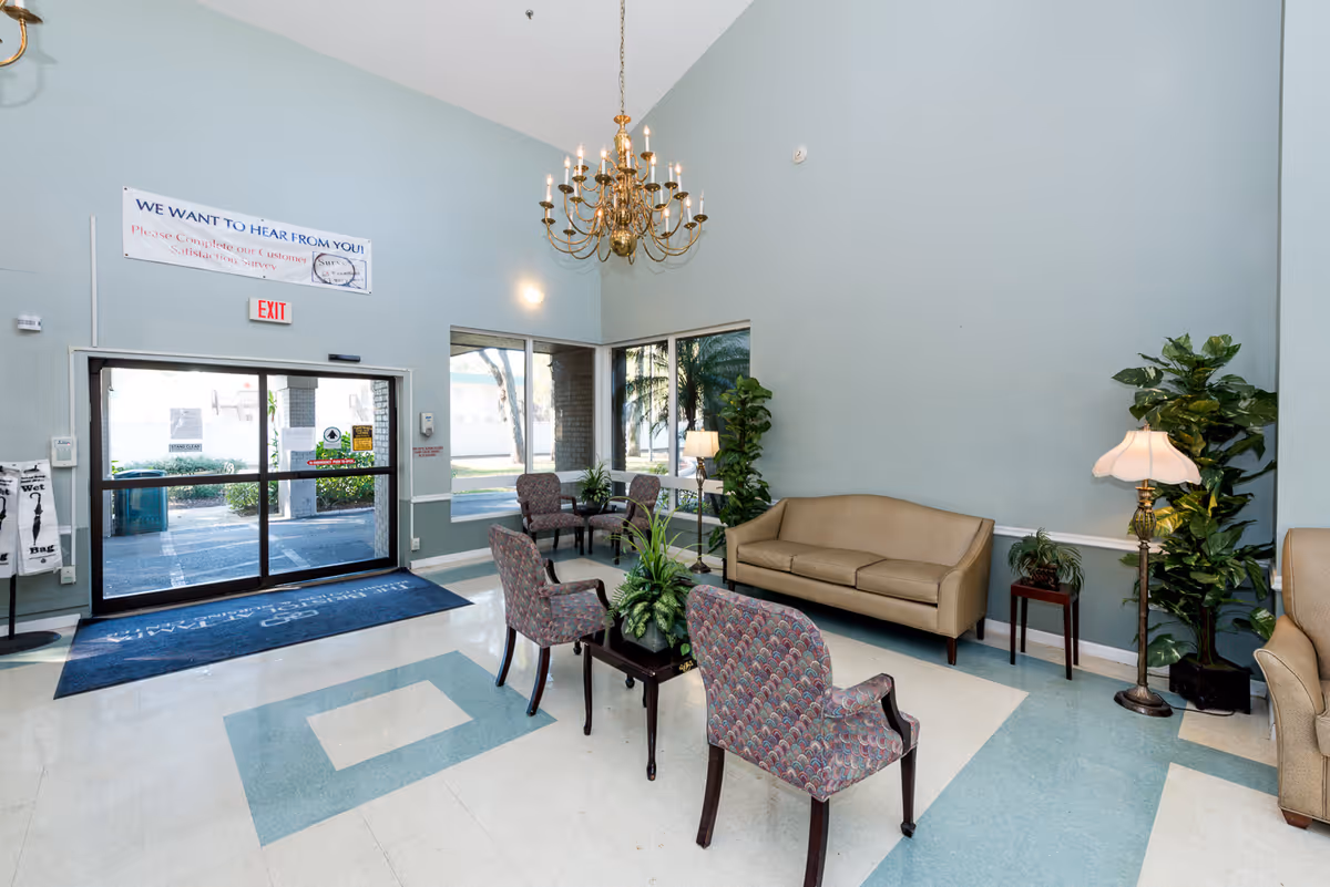 A spacious lobby area with light blue walls and a high ceiling featuring a large chandelier. The room has a beige sofa, patterned armchairs, several potted plants, and floor lamps. There is a glass double door entrance with an exit sign above it and large windows letting in natural light. A sign above the door reads 'WE WANT TO HEAR FROM YOU! Please Complete our Customer Satisfaction Survey.'